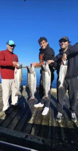 Three anglers on wooden dock displaying four chinook salmon under clear blue sky over calm water.