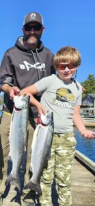 Man and boy displaying two coho salmon on wooden dock with blue sky and water background