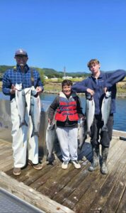 Three anglers on wooden dock displaying six coho salmon with coastal bay and mountains behind them
