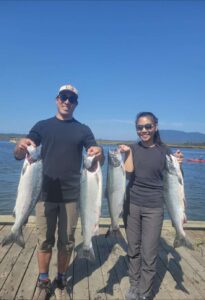 Two anglers on wooden dock displaying four coho salmon under clear blue sky with water and mountains behind.
