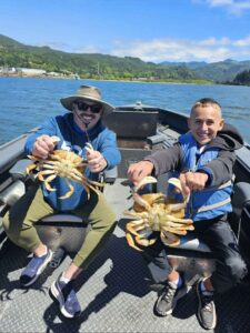 Two anglers in blue life jackets display two large Dungeness crabs on a boat with forested mountains behind.