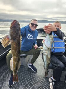 Two anglers in boat displaying large lingcod catches under overcast skies on calm water with mountains visible.