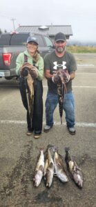 Two anglers holding lingcod with six additional lingcod displayed on wet pavement in parking area
