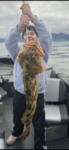 Angler holding large lingcod vertically on boat with mountains and water in background