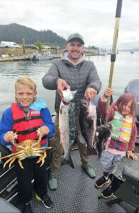 Adult and two children on boat displaying multiple fish including Dungeness crab and salmon on overcast day