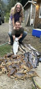 Two people display mixed catch of Dungeness crabs, salmon, and rockfish on gravel driveway